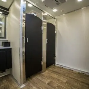 Interior of a modern mobile bathroom trailer featuring two closed stall doors, a sink with a black countertop, mirrors, and wood-like flooring