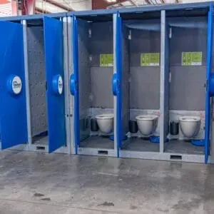 A row of open blue portable toilets, part of the USS Hydroflow System, in a large industrial space, each featuring a white toilet and hand sanitizer
