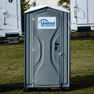 A standard portable toilet outdoors with white portable restroom trailers visible in the background, set on a grassy area