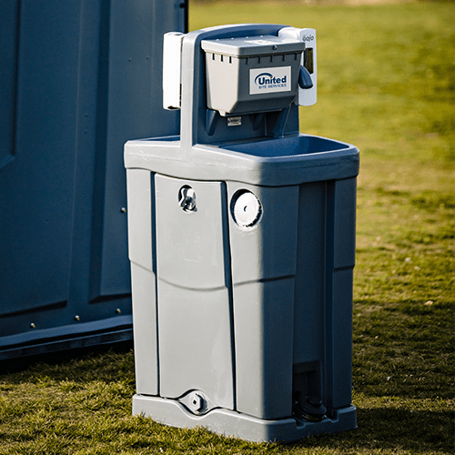 A portable gray handwashing station with a paper towel dispenser and a soap dispenser, set on a grassy area beside a blue portable toilet